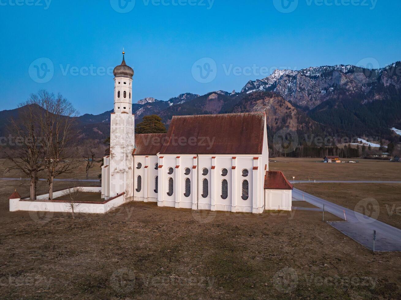 St. Coloman Church stands in an open field with bare trees, framed by the Bavarian Alps and a clear blue sky. Snow capped peaks are visible in the distance. photo