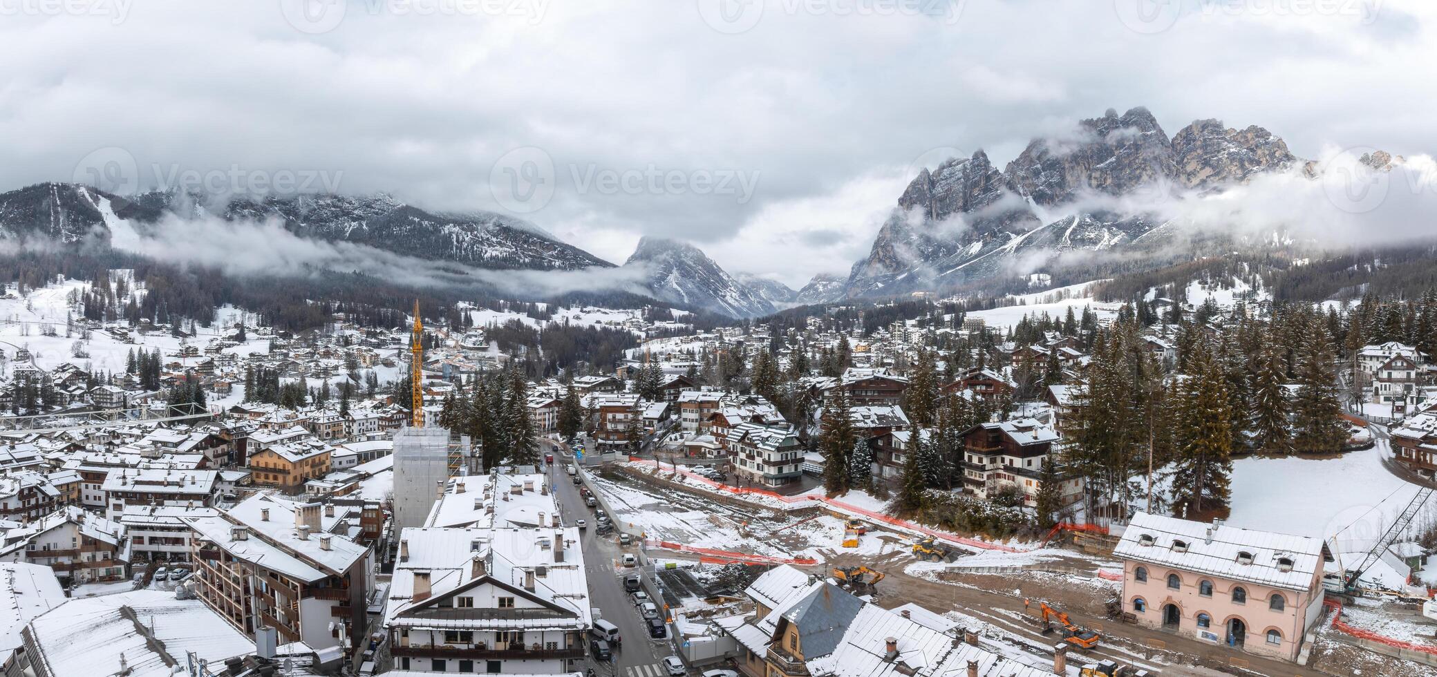 Aerial view of Cortina d'Ampezzo in Italy, featuring snow covered alpine buildings, a church spire, and misty Dolomite mountains in the background. photo