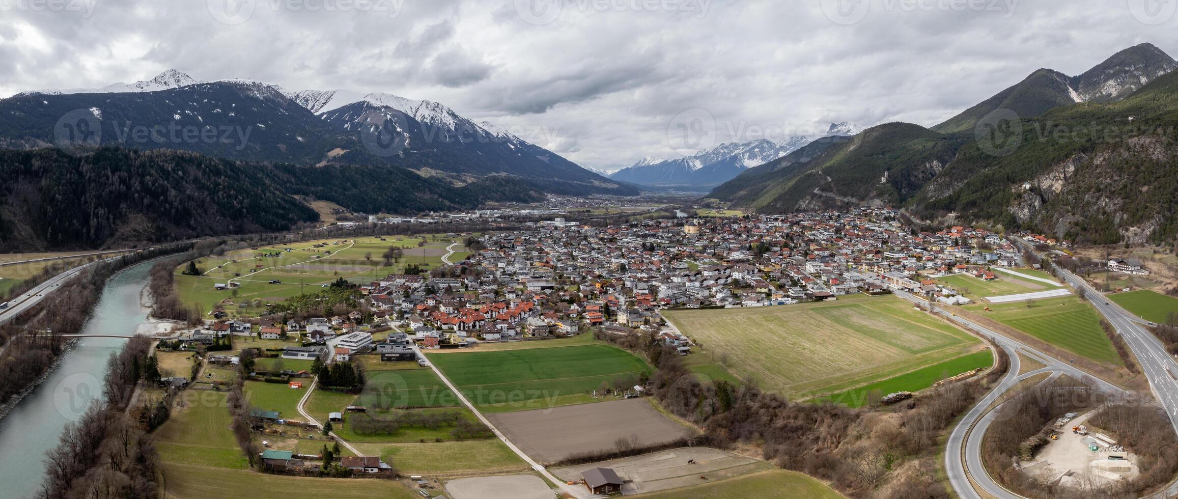 A small Austrian town surrounded by green fields, forested hills, and dramatic snow capped mountains. A river and bridge run along its edge. photo