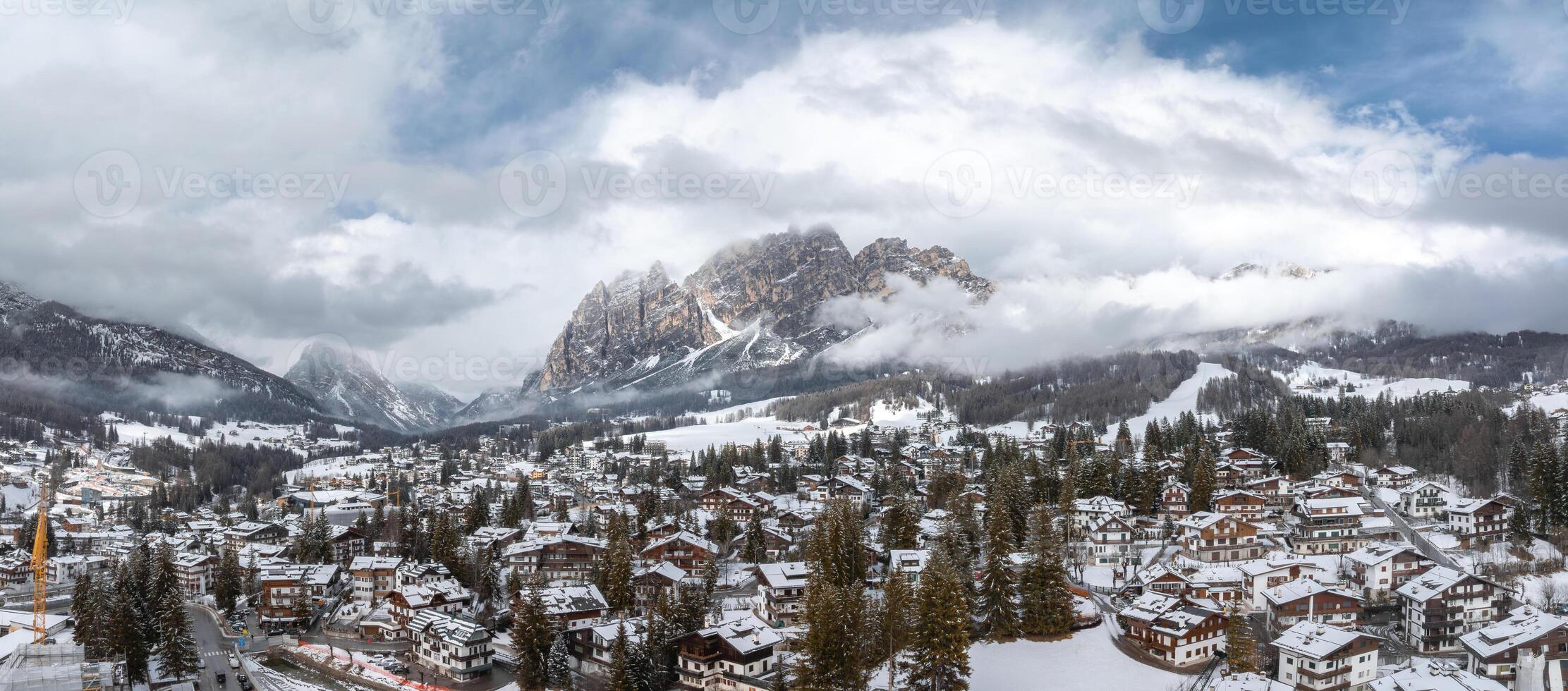 Aerial view of Cortina d'Ampezzo in Italy with snow covered rooftops, evergreen trees, Dolomites peaks, low clouds, and a visible construction crane. photo