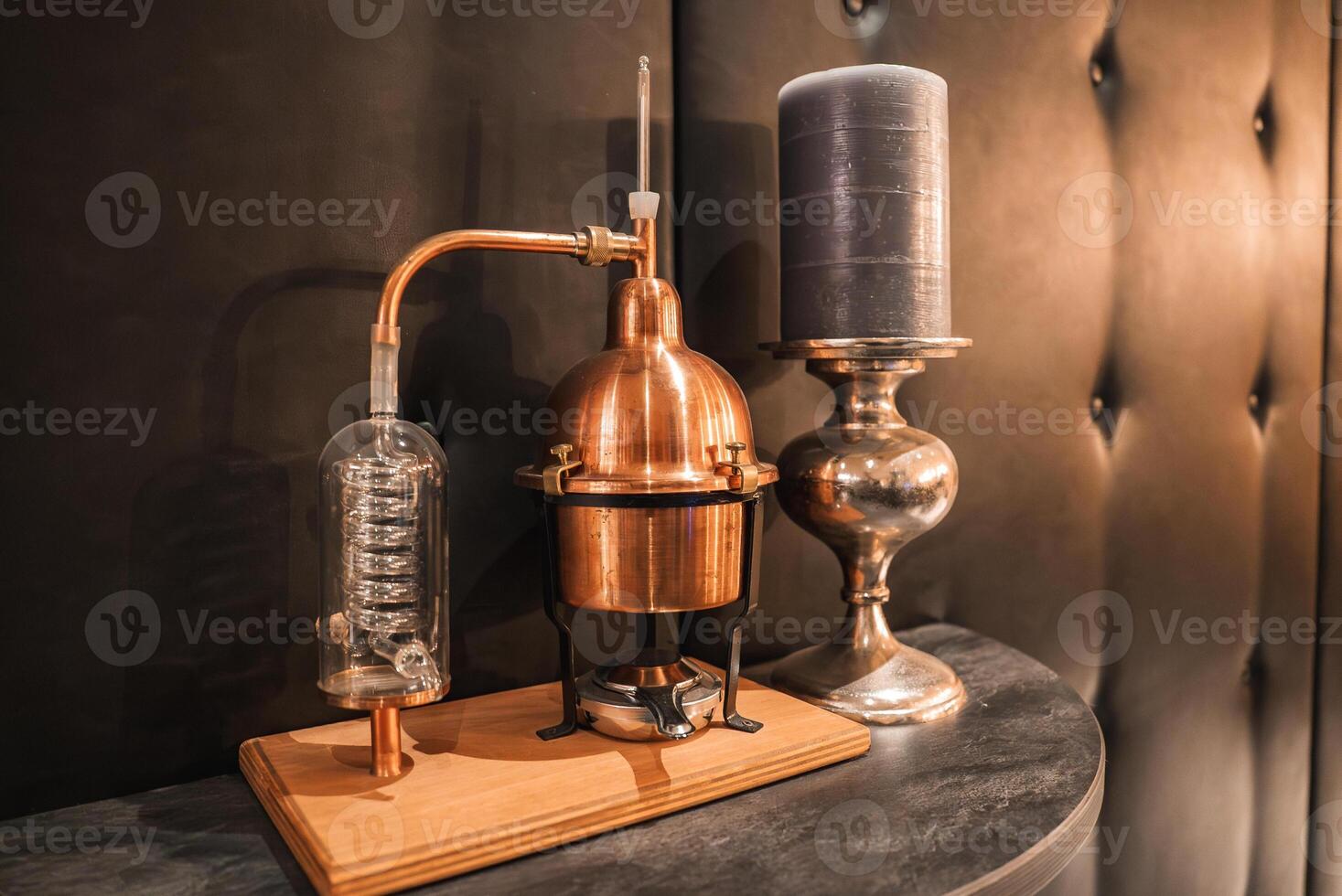 A copper distillation apparatus with glass coil on a wooden base, paired with a dark gray candle in a silver holder against a tufted leather backdrop. photo