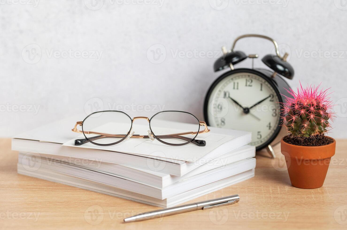 Eyeglasses resting on stack of notebooks with pen, alarm clock, and cactus photo