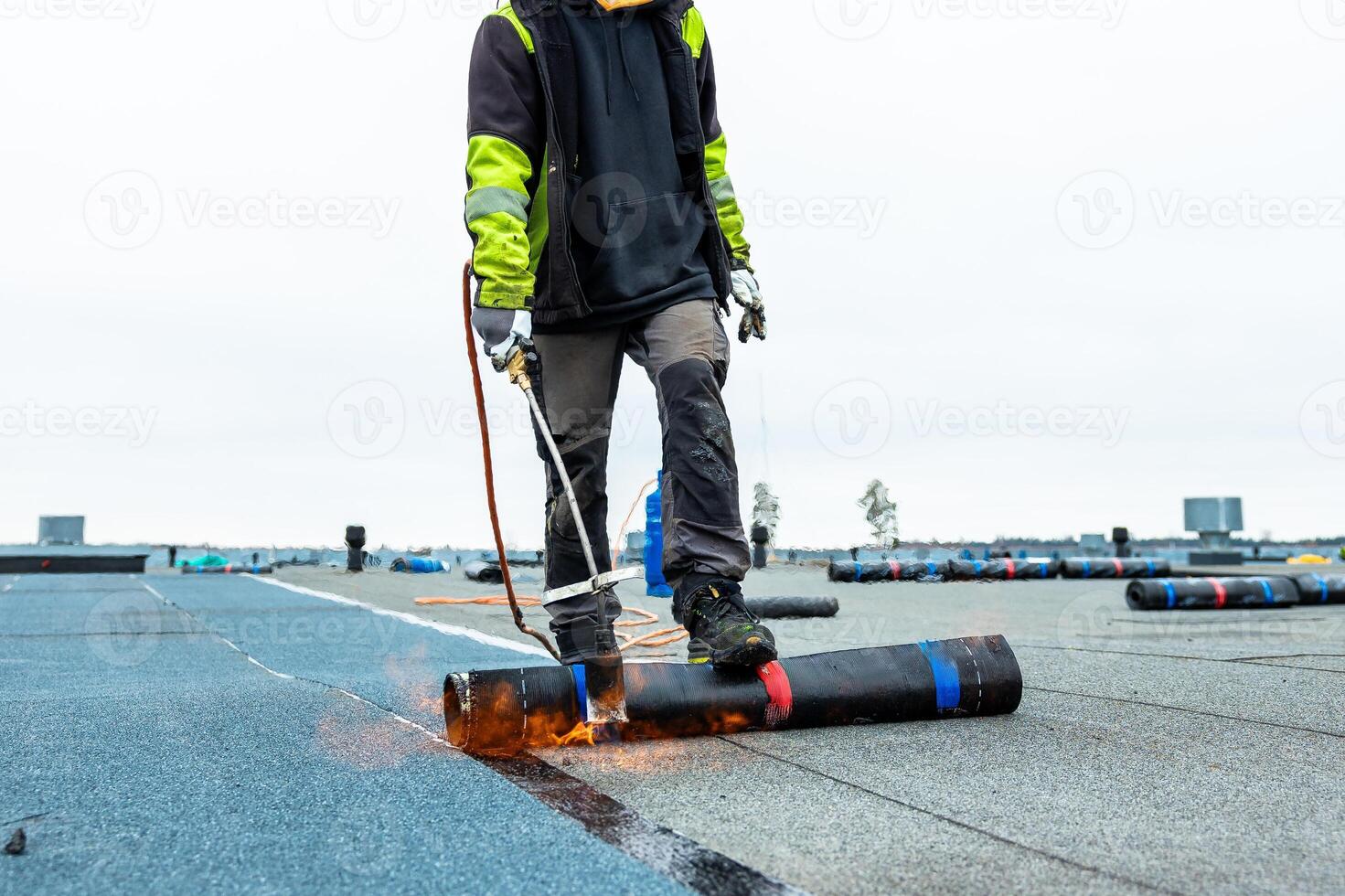 A worker in protective gear uses a torch to apply roofing material on a flat rooftop. Rolls of material and distant structures are visible under a cloudy sky. photo