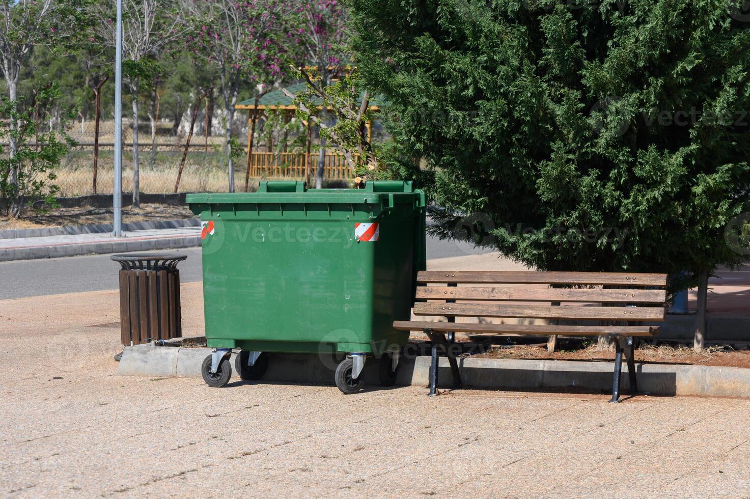 Green dumpster and wooden bench in a serene park setting during a sunny afternoon near blooming trees photo