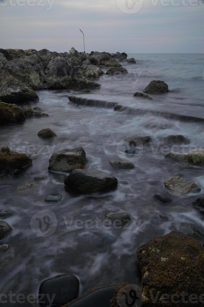 Gentle waves caress the rocky shore under a soft, cloudy sky at dusk by the tranquil coastline photo