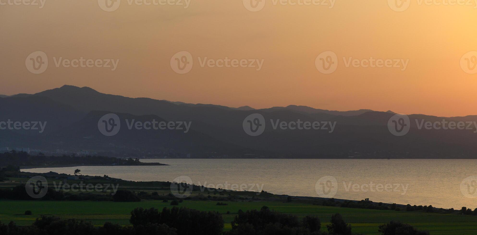 Calm waters reflect the golden hues of sunset over distant mountains and lush fields in a tranquil landscape photo
