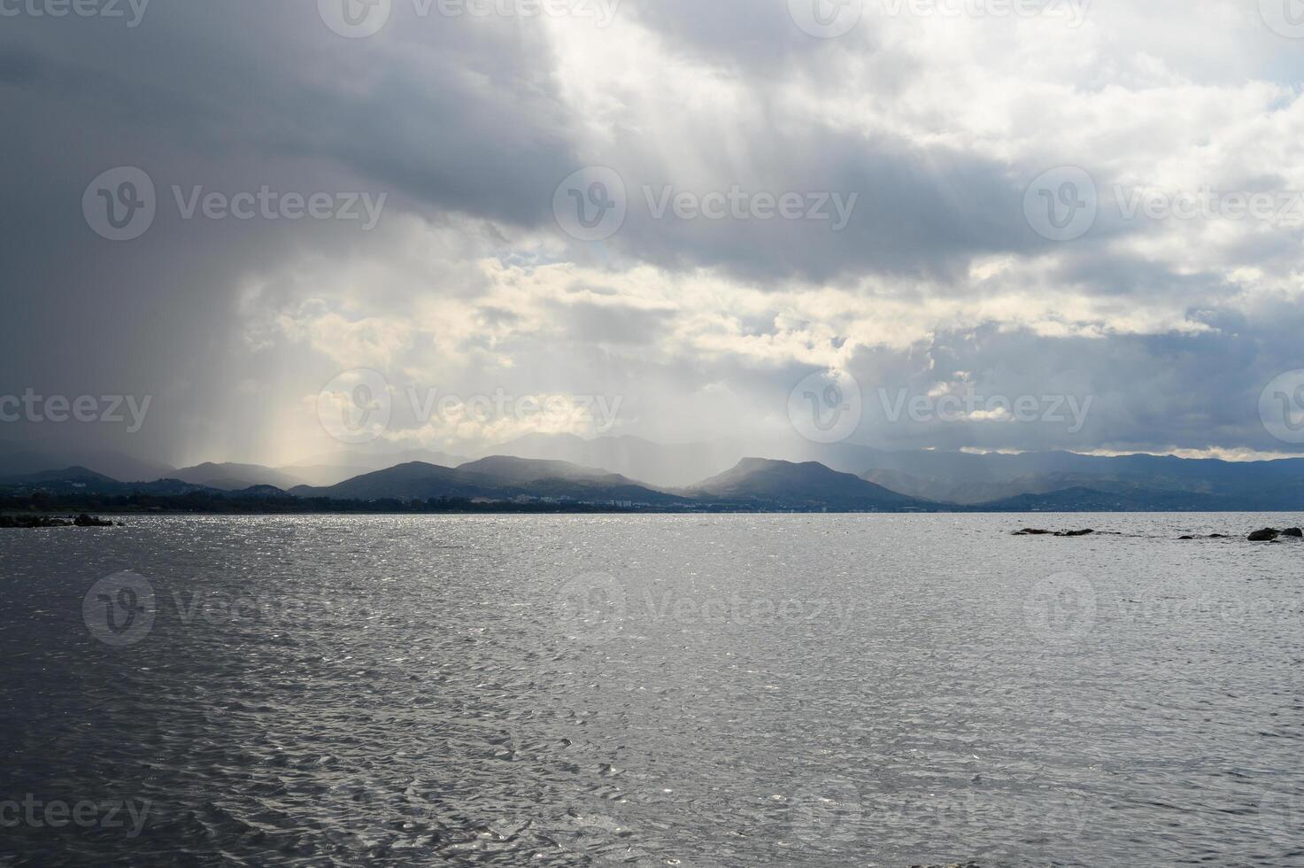 Dramatic clouds cast shadows over tranquil waters near distant mountains during a cloudy afternoon photo