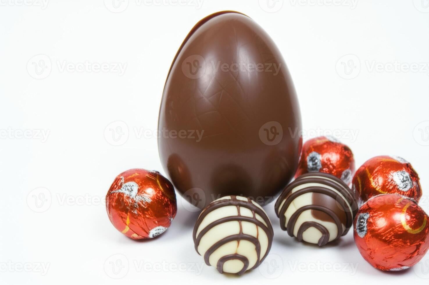 A large chocolate egg and several chocolates are placed on a white background. Three round candies are wrapped in red foil, and the other two have a chocolate striped shell, creating a sweet treat. photo