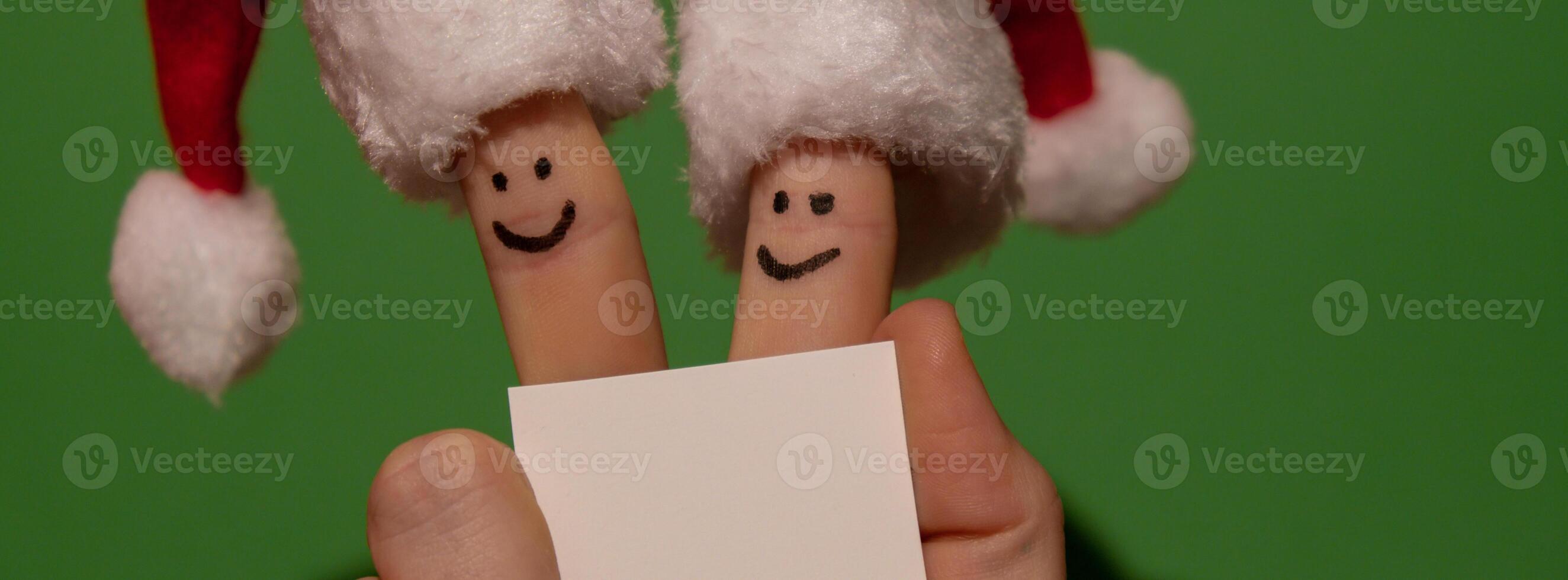 Fingers dressed in Santa-Claus red-white hats. Two happy finger smileys faces holding empty paper note for your text copy space against green background. Happy family celebrating concept for Christmas or New Years day. Finger couple Advertisement photo