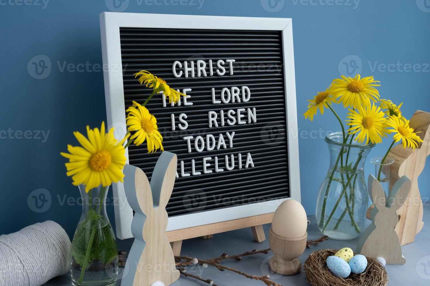 Phrase CHRIST THE LORD IS RISEN TODAY ALLELUIA traditional greeting to christian holiday. Festive mood composition with bunnies and yellow flowers on pastel blue background. Sustainable natural materials decorations photo