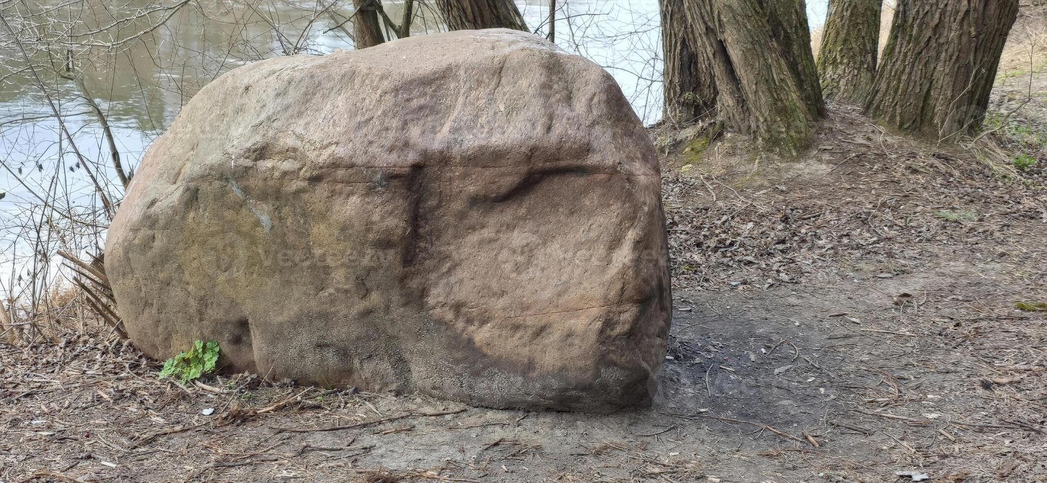 A large boulder resting by a riverbank, illustrating the natural beauty and permanence of geological formations in outdoor settings. photo