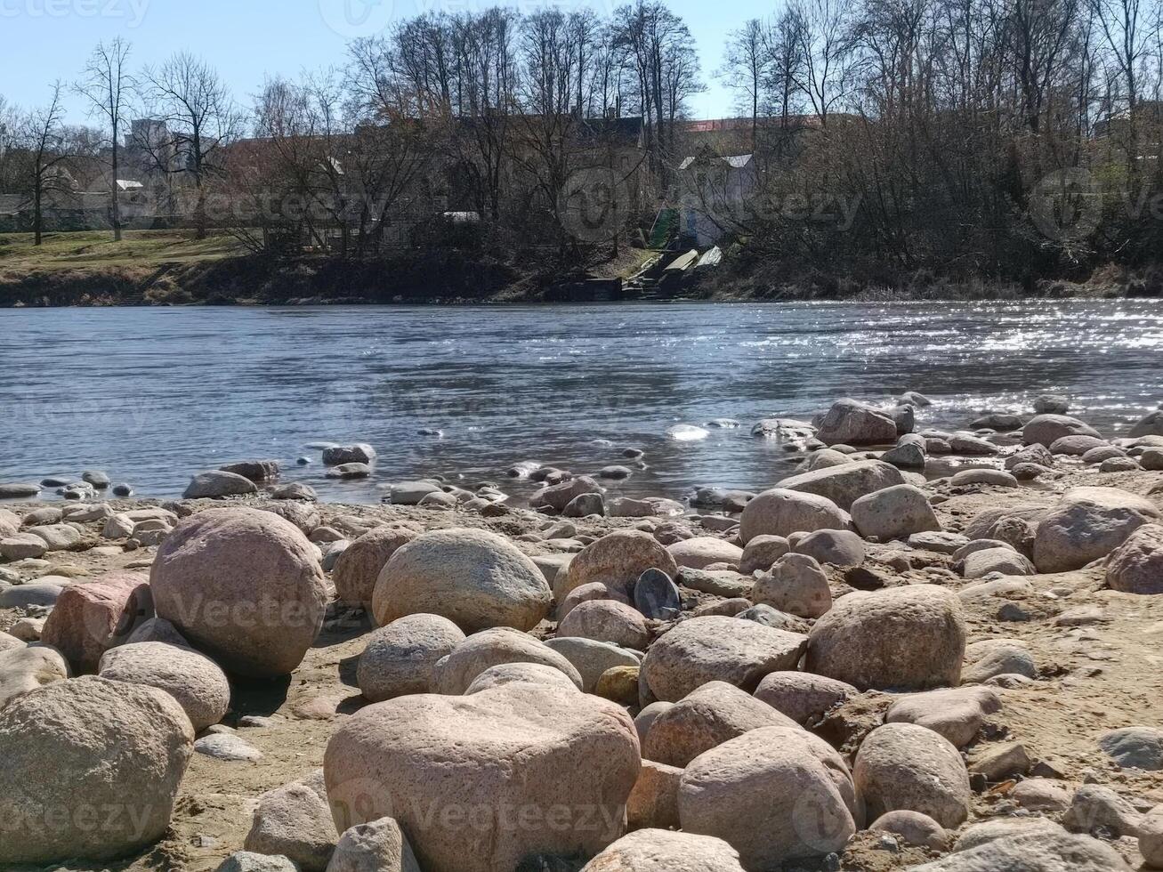 Smooth, rounded stones line the riverside, with a calm river and trees in the background, creating a serene outdoor scene. photo