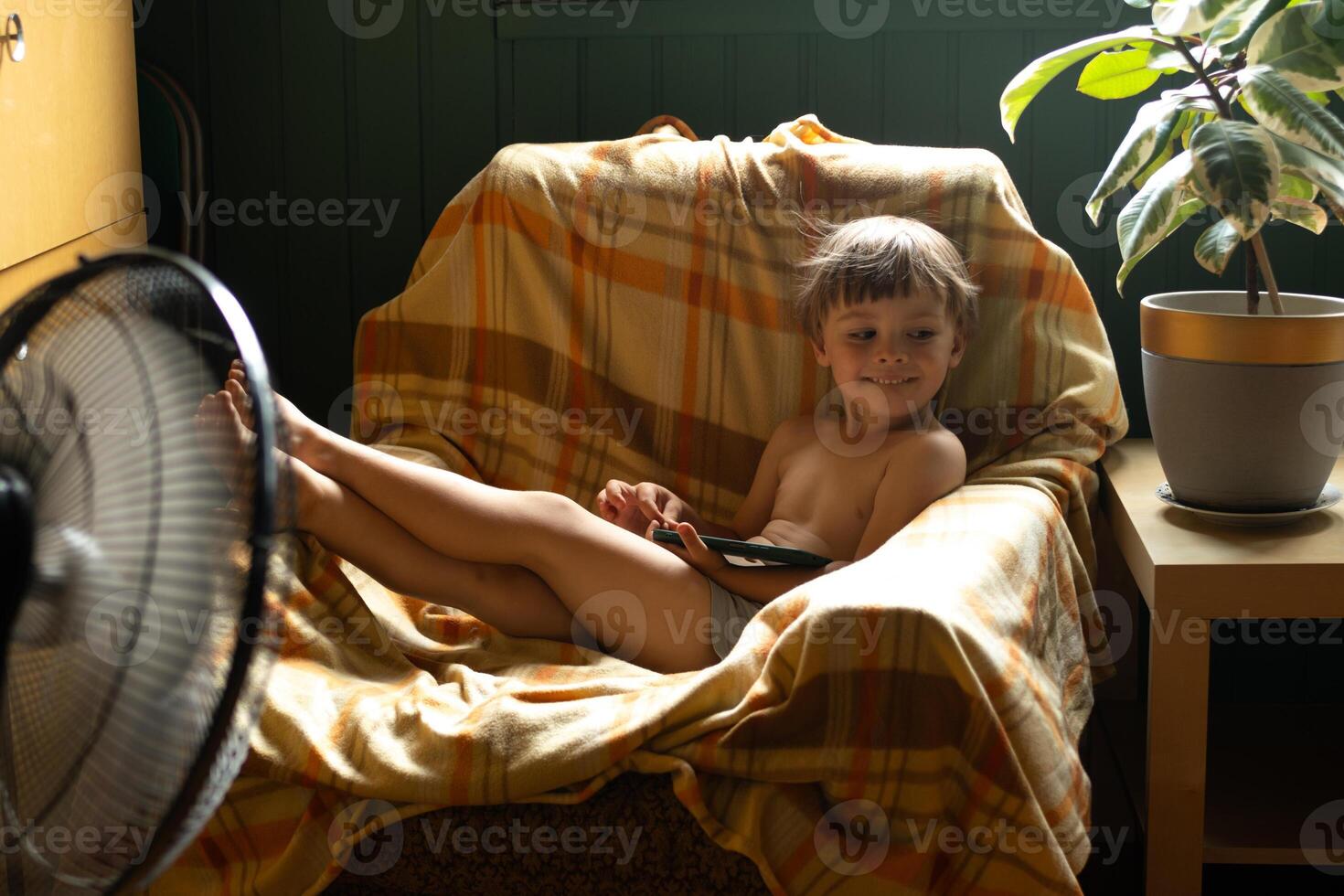 Summer vibes, hot weather at home. Small child sitting in chair with smartphone and enjoying breeze from fan. photo