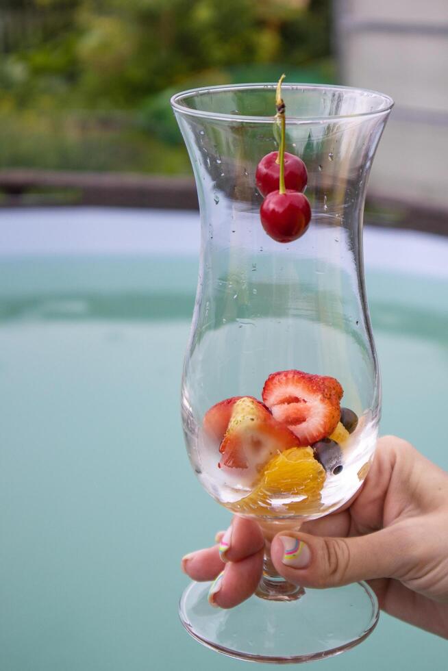 A summer cocktail with strawberries, cherries and foamy champagne in a woman's hand, against a blue pool backdrop. Party concept, vertical photo