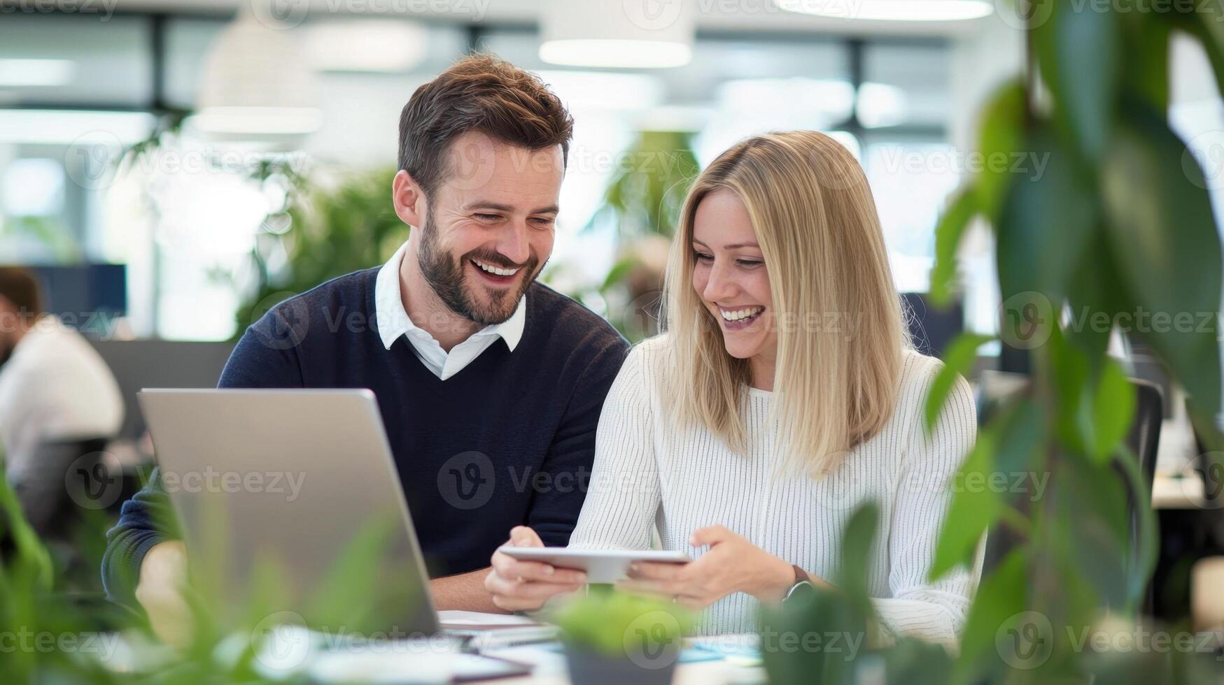 man and woman share a laugh while working at and desks in a contemporary office. They look at a tablet together, creating a collaborative their friendly work environment photo