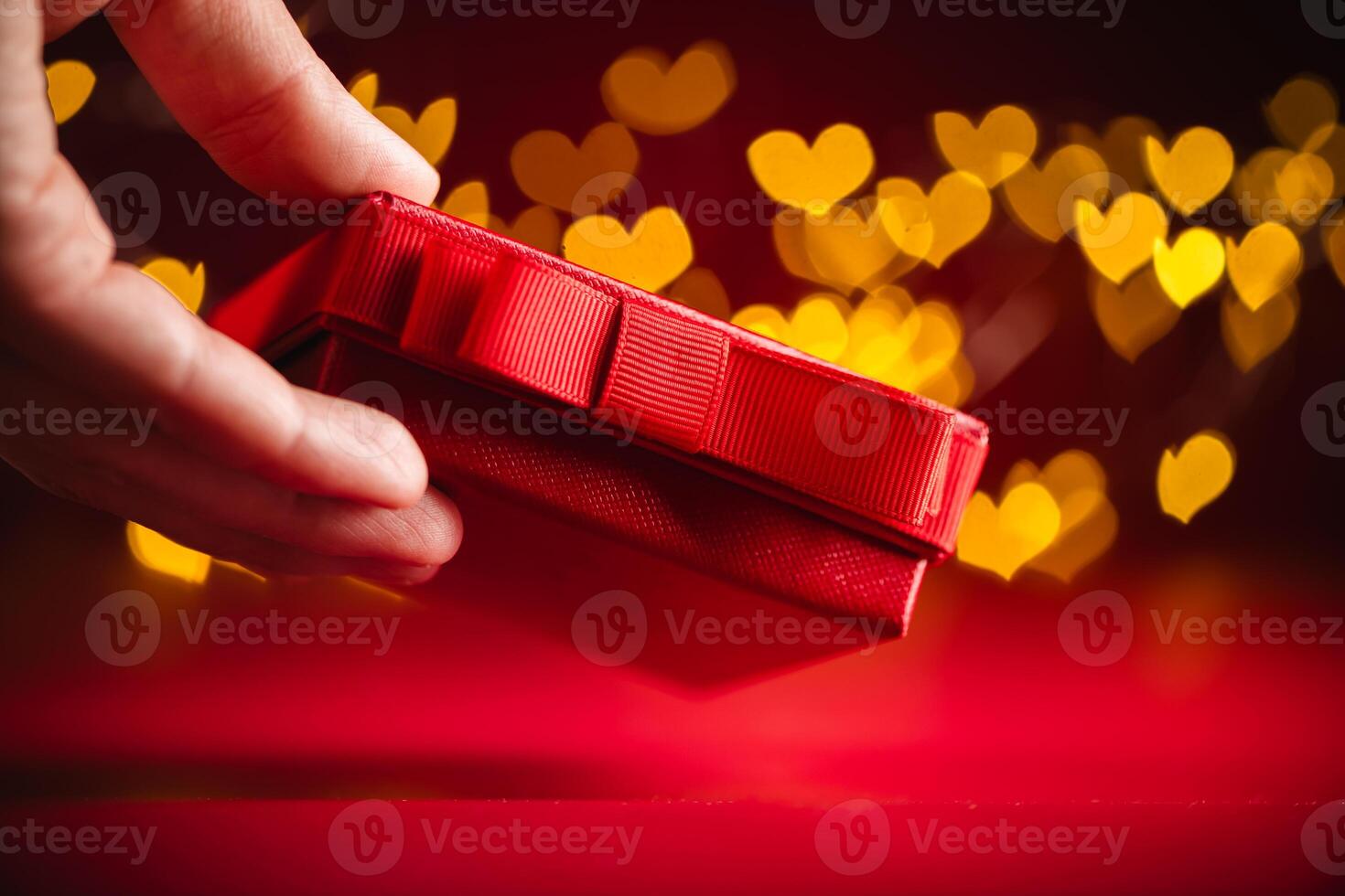 Hand gracefully presenting a red gift box adorned with a ribbon, set against a backdrop of glowing, heart-shaped bokeh lights, symbolizing love and celebration on valentine's day photo