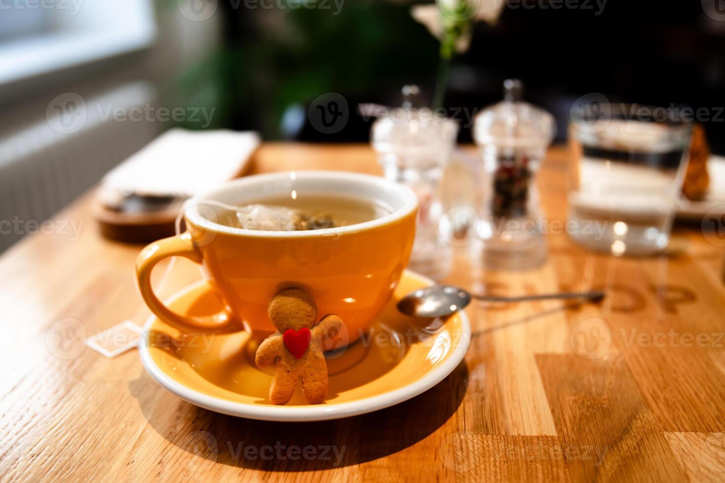 Gingerbread man with a red heart is hugging a yellow cup of tea, sitting on a saucer with a spoon, on a wooden cafe table, creating a cozy and festive atmosphere photo