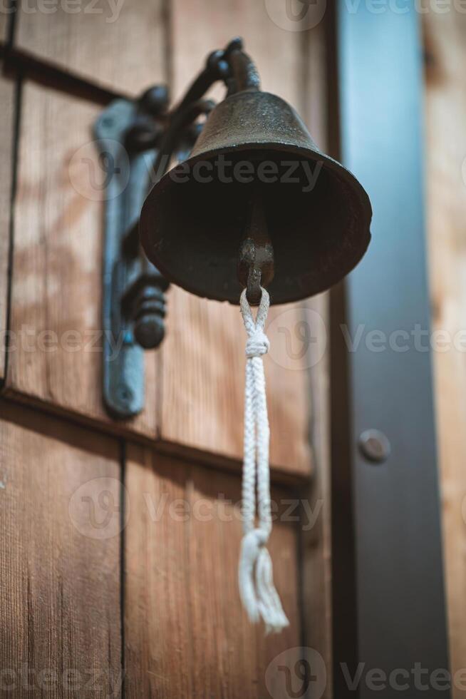 Hanging on a rustic wooden door, an old metal bell with a white rope awaits ringing, ready to announce arrivals or signal important events with its classic sound photo