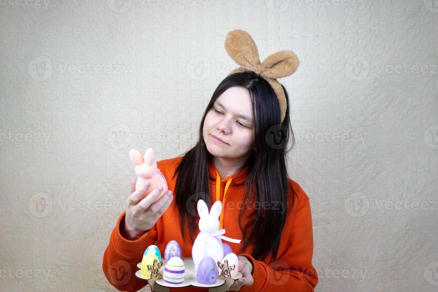 A pretty girl in casual clothes with brown bunny ears, holding a stand with Easter eggs and bunnies, looks at a rabbit . photo