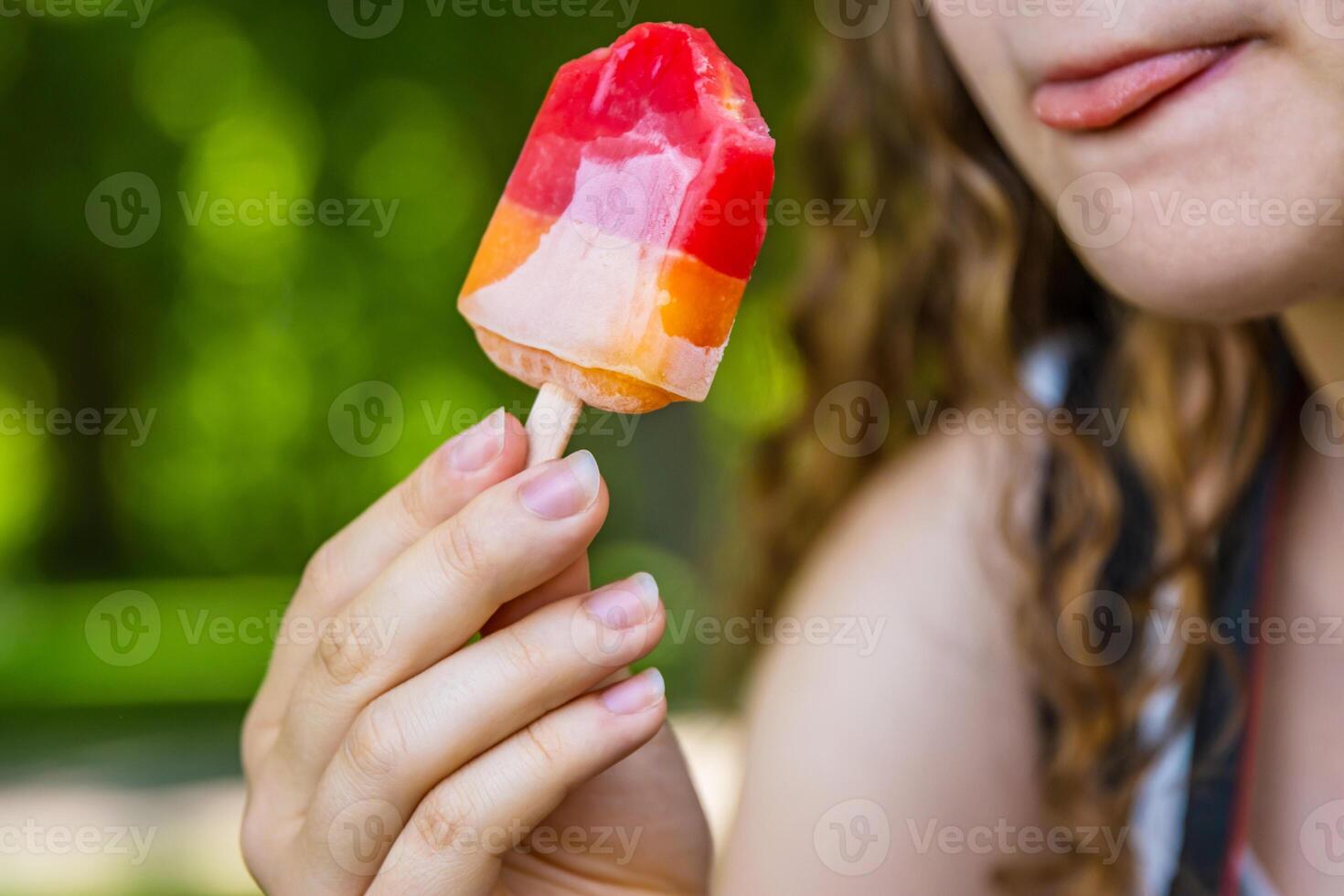 Crop image of the teenager girl eating ice cream. Young lady is holding colorful melted bitten fruit ice cream popsicle on a hot summer day, Selective focus, Burred green background. Copy space 02 photo