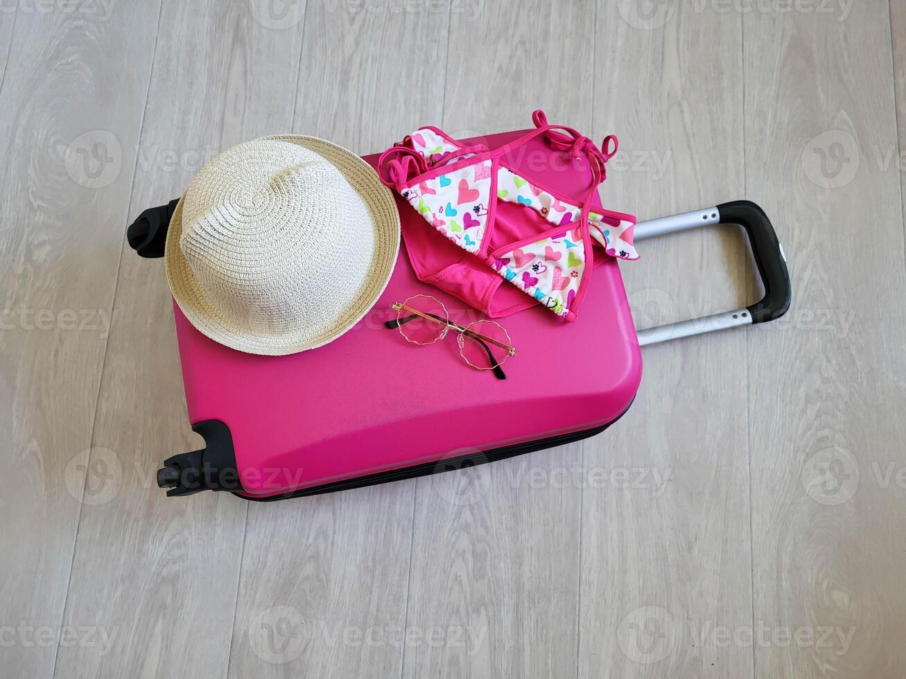 On the pink children's suitcase are a children's swimsuit, hat and sunglasses. A suitcase on wheels with a handle is lying on the floor. photo
