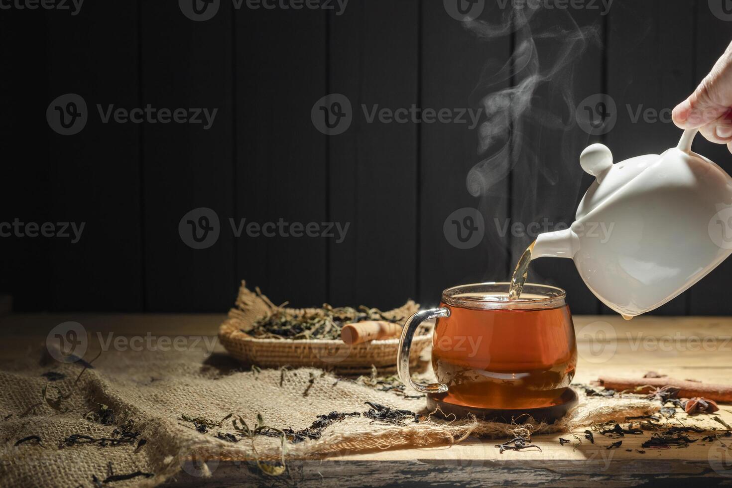 Pour hot tea into a cup, a glass of herbal tea is placed on a wooden table, a white teapot in a rustic style. photo
