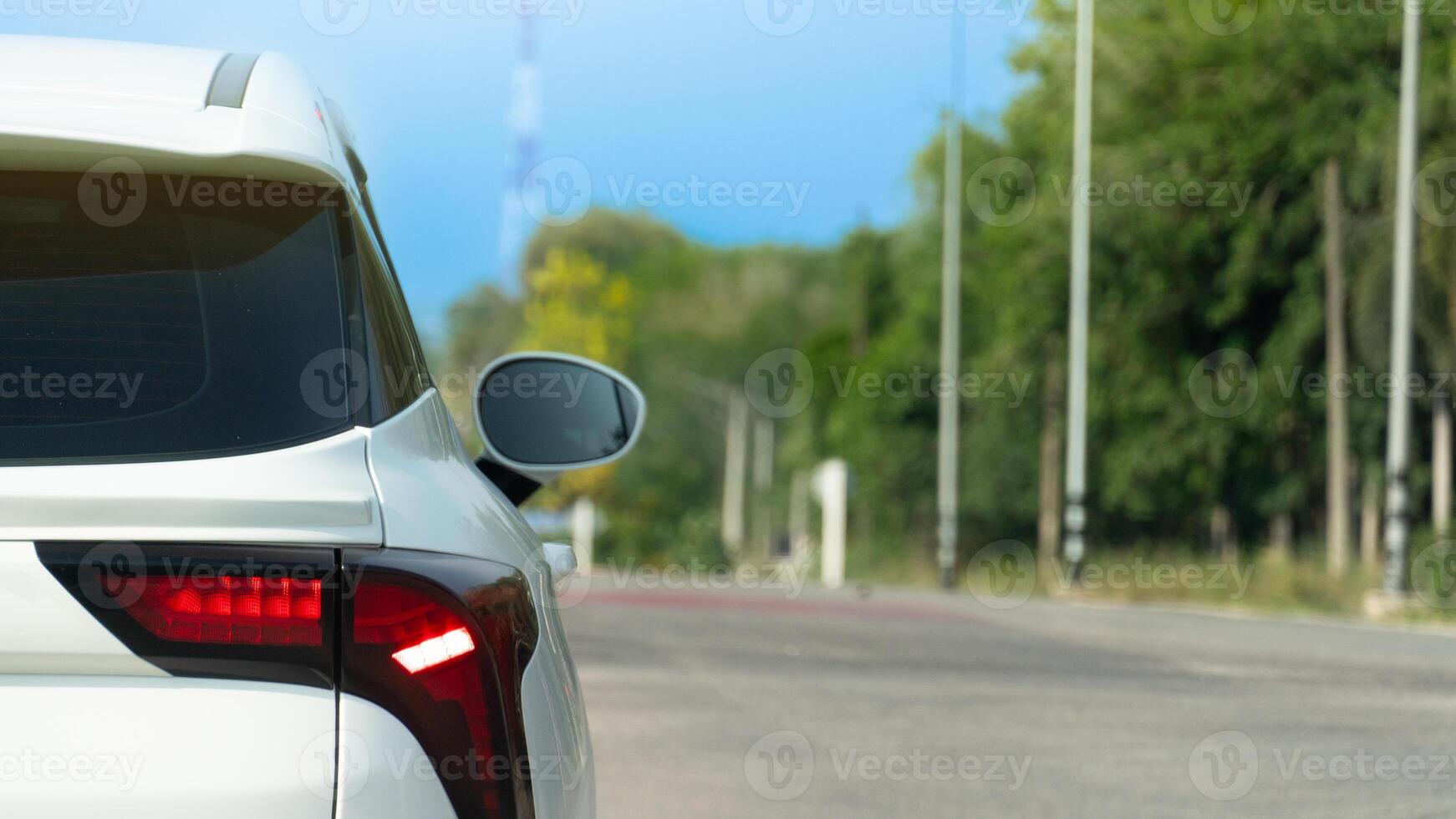 Rear side of white car with trun on brake light. Clear road in bright light. Background of pole and green forest under blue sky. photo