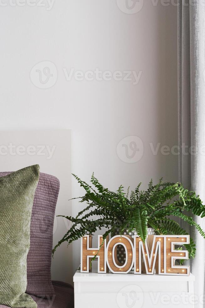 Bedroom interior, bedside table with 'Home' sign, potted fern plant and bed with green and purple pillows. Cozy home concept photo