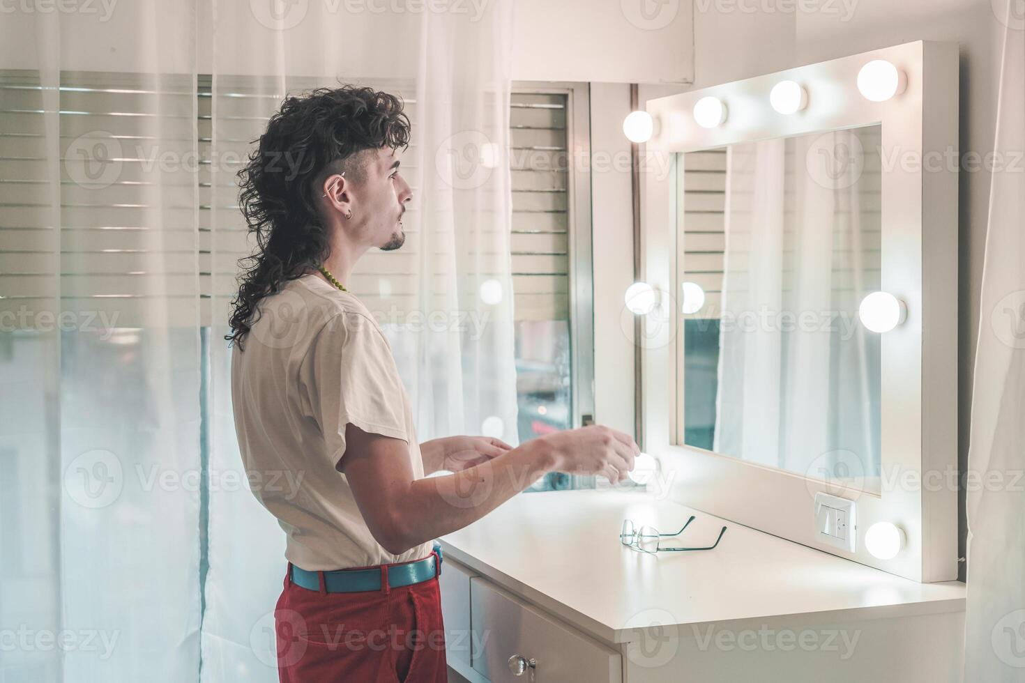 Young man with mullet hairstyle preparing in front of mirror with lights getting ready in the morning at home photo