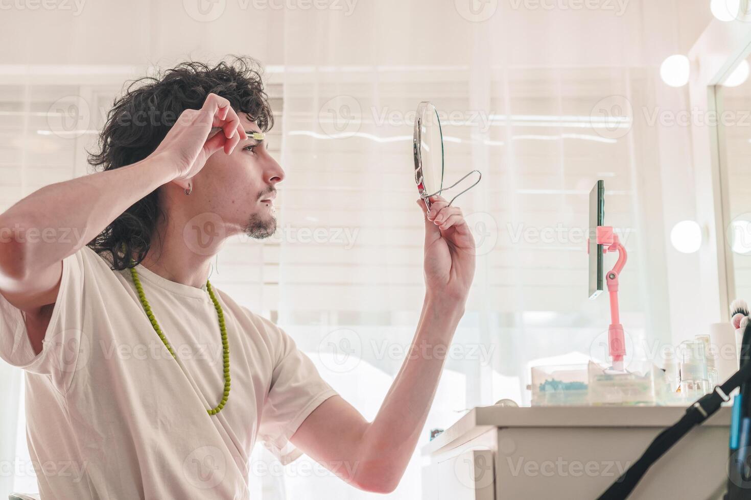 Young man plucking eyebrows looking at a small mirror and recording with smartphone photo