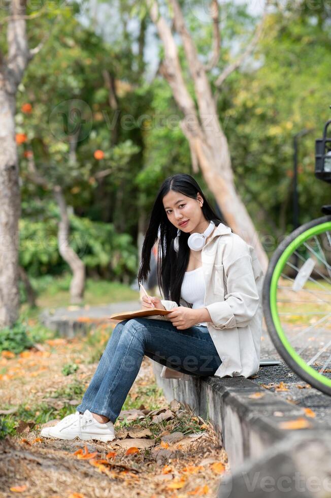 Asian woman is looking straight into camera while writing on her notebook, sitting on park sidewalk. photo