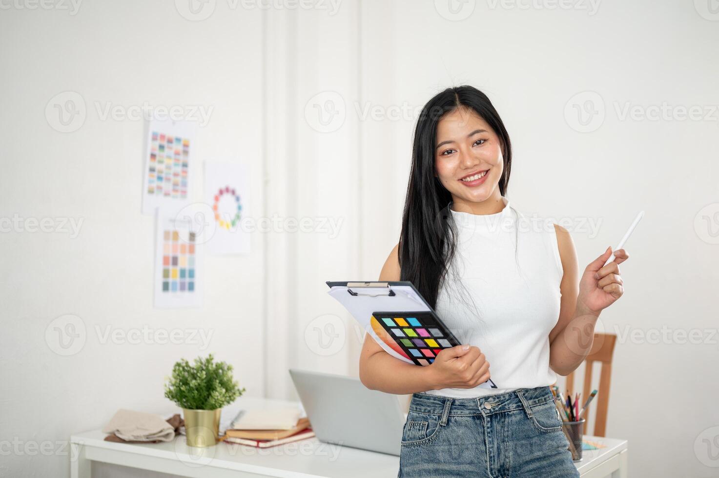 A smiling asian woman is carrying a clipboard and a stylus in front of her work desk. Designer work. photo