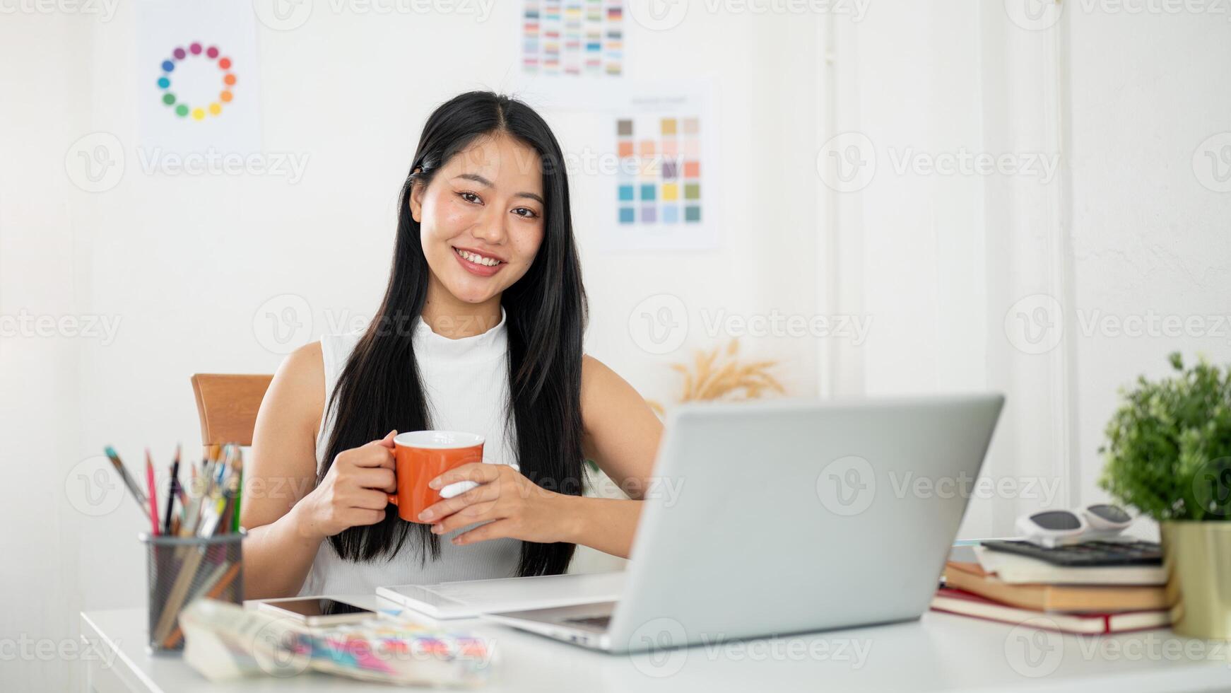 A smiling asian woman is holding a mug with both hands, sitting in front of a laptop on working desk photo