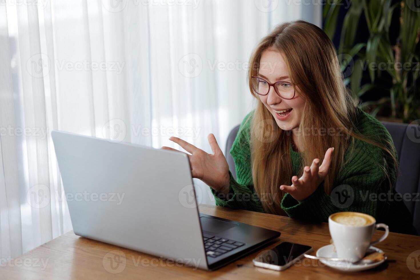 Happy woman with glasses celebrates while using laptop during online conversation in cafe. photo