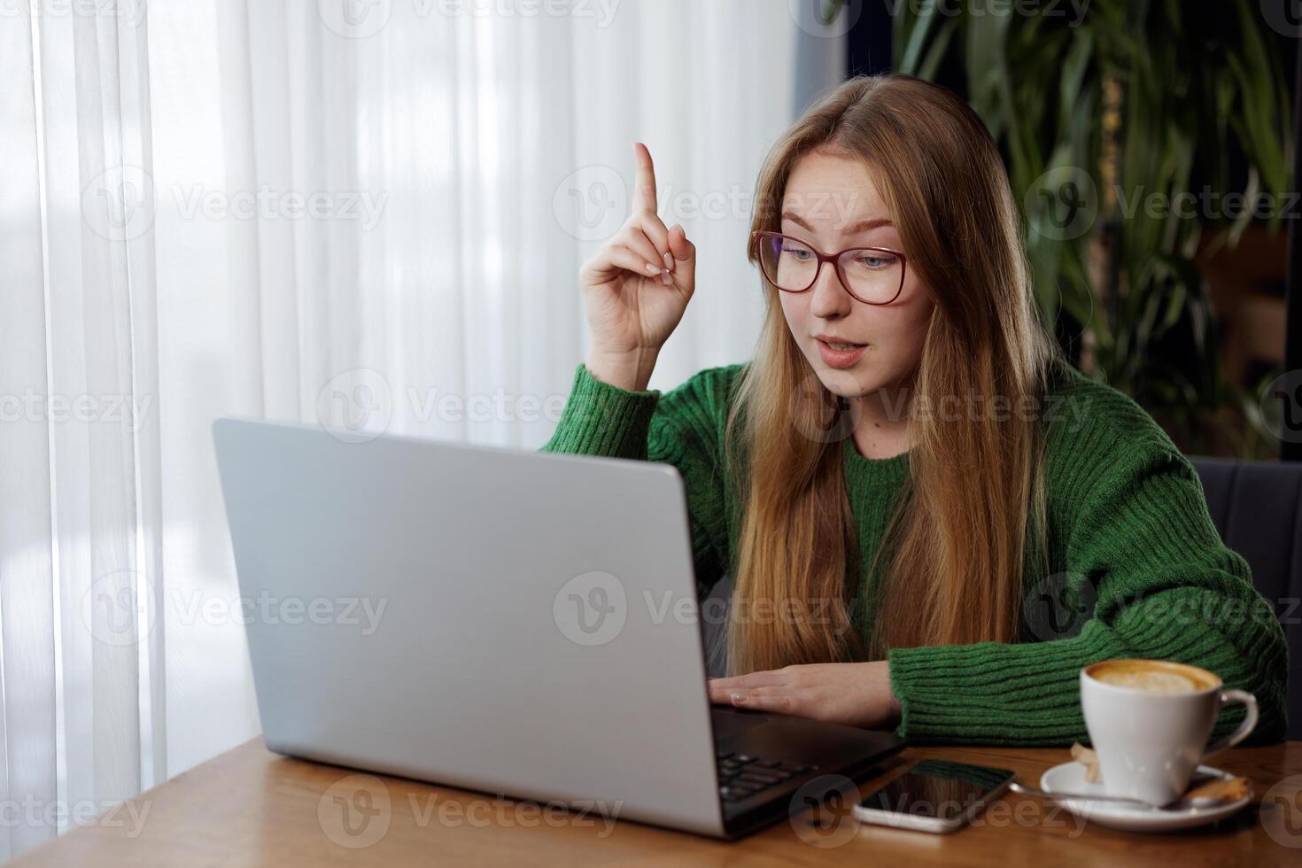 Woman in glasses raises finger with new idea while having a call on laptop at a cafe. photo
