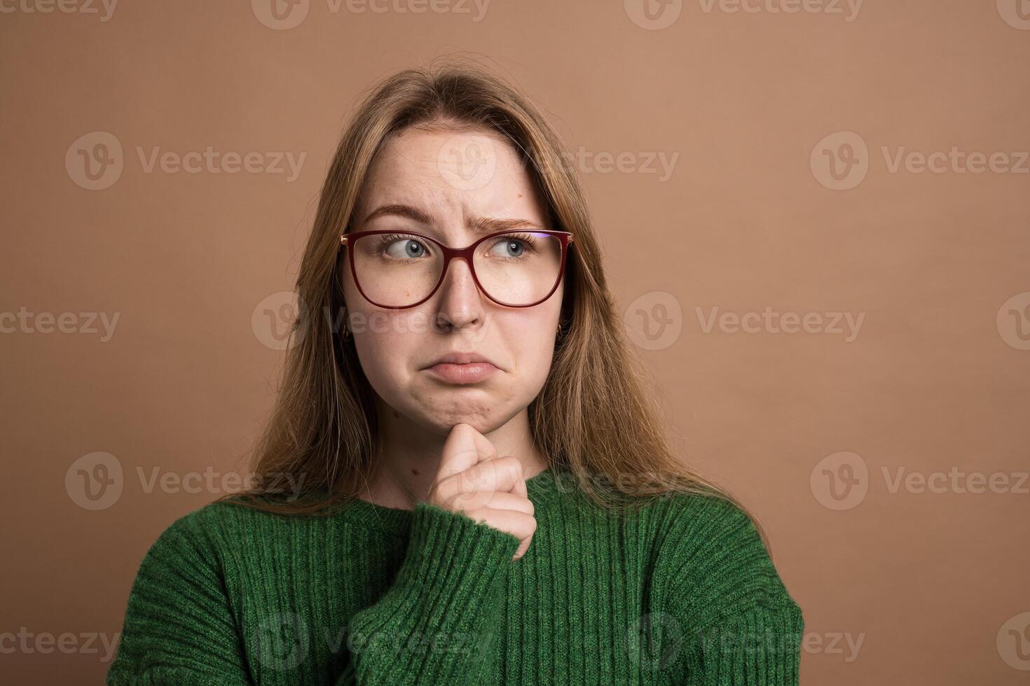 Pensive woman in green sweater looks uncertain or confused, touching chin and raising eyebrows. photo