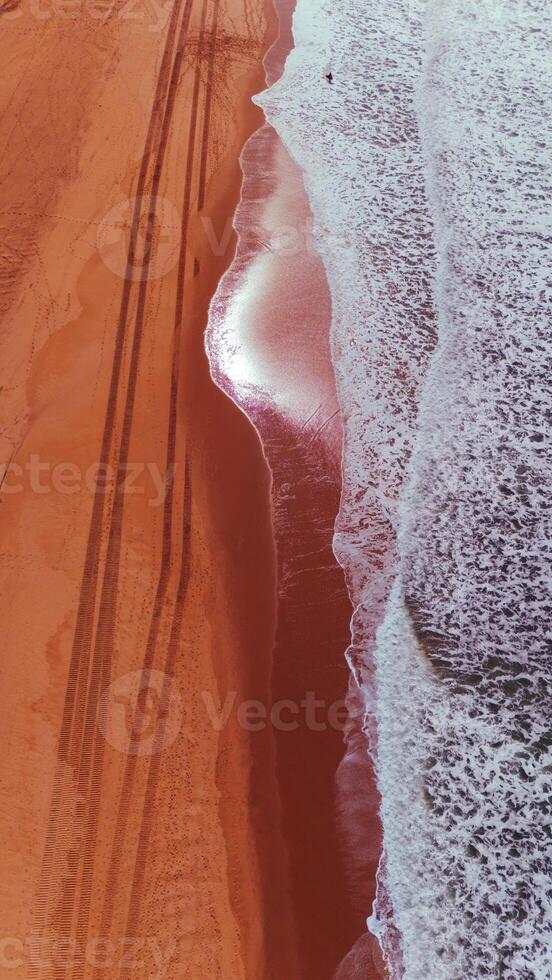Overhead Drone View of Ocean Waves and Tire Tracks on Red-Toned Beach photo
