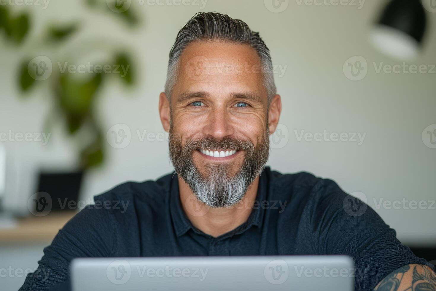 A white man with a beard and detailed arm tattoos sits at a desk, smiling while focused on his laptop in a well-lit office photo