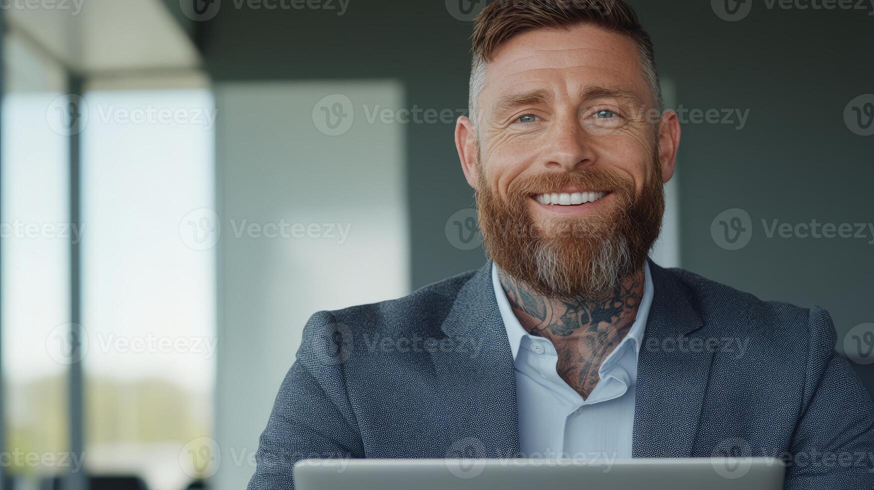 A white man with a well-groomed beard and arm tattoos is smiling while working on a laptop at a sleek office space photo