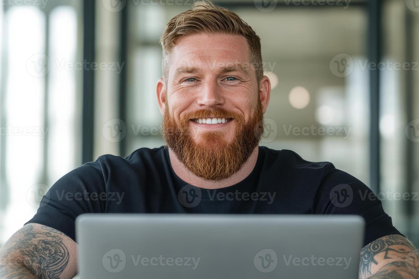 A white man with a beard and tattoos sits in a contemporary workspace, smiling while engaged with his laptop, creating a vibrant atmosphere photo