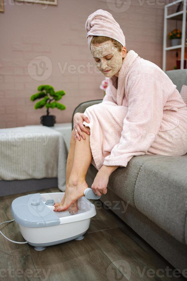 Woman enjoying a foot spa treatment with a face mask at home photo