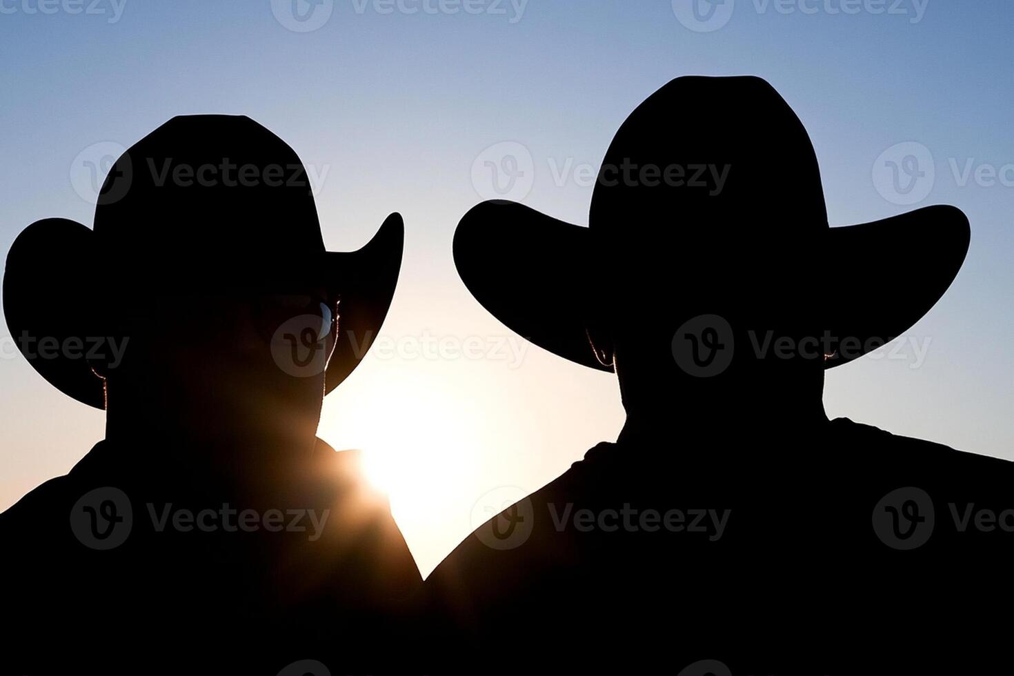 Two men wearing cowboy hats stand in silhouette against the sun photo
