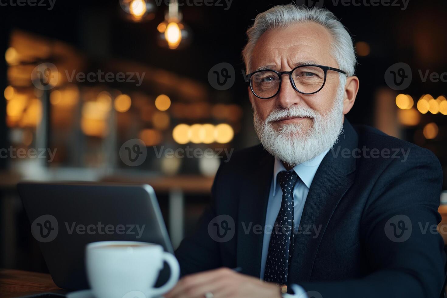 An older man with glasses and a beard sitting at a table with a laptop photo