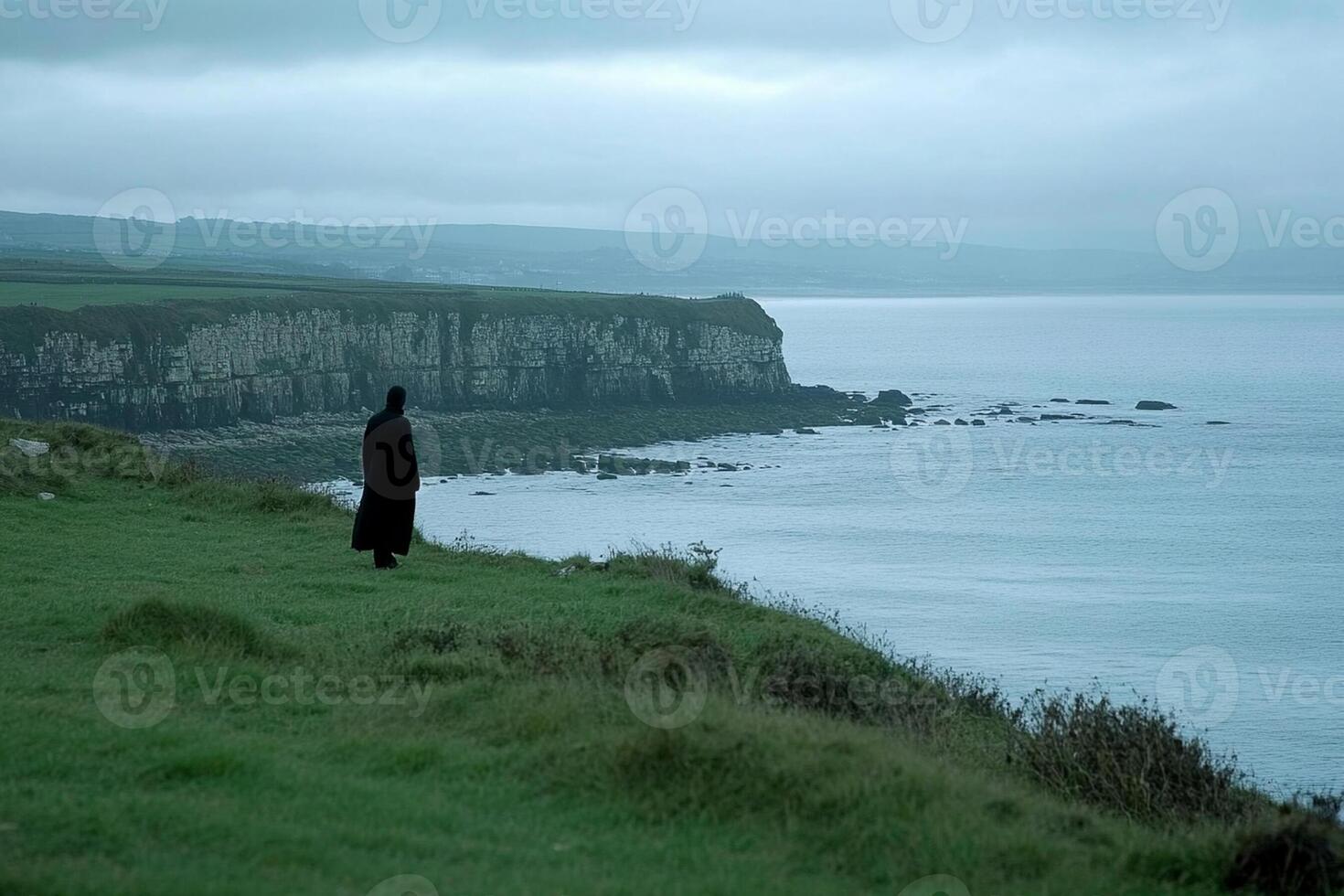 A person in a black cloak standing on a hill overlooking the ocean photo