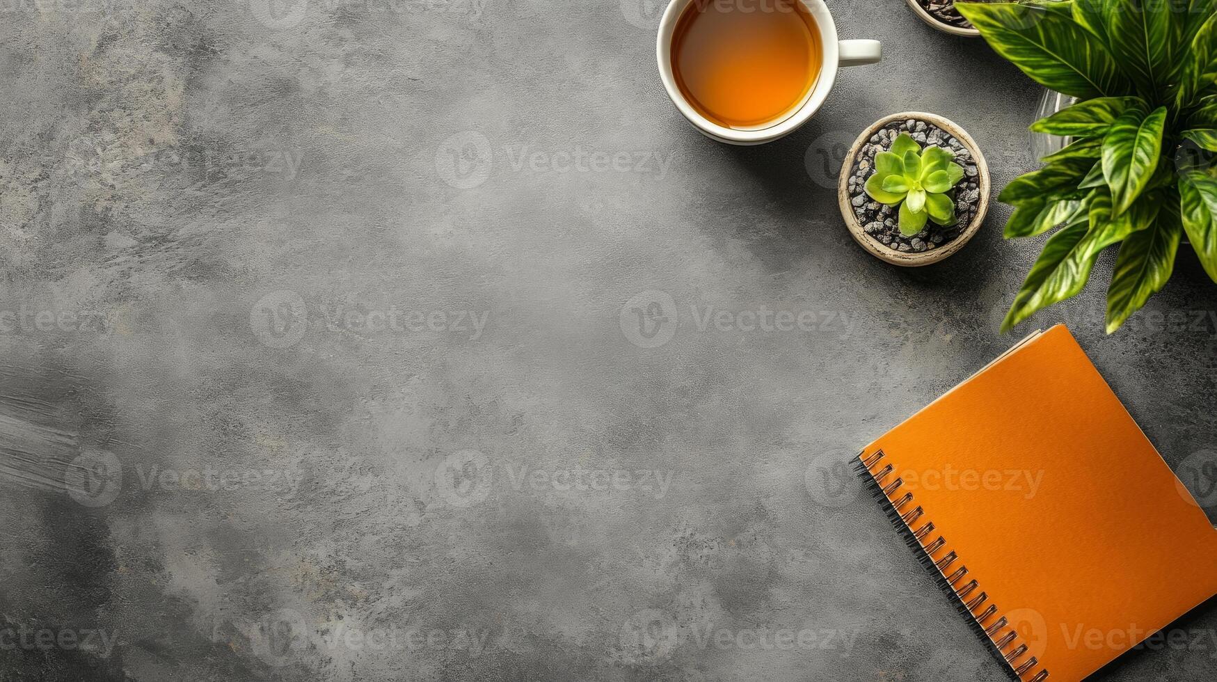 A serene workspace features a cup of tea, two small potted plants, and a vibrant notepad, arranged neatly on a grey textured surface photo