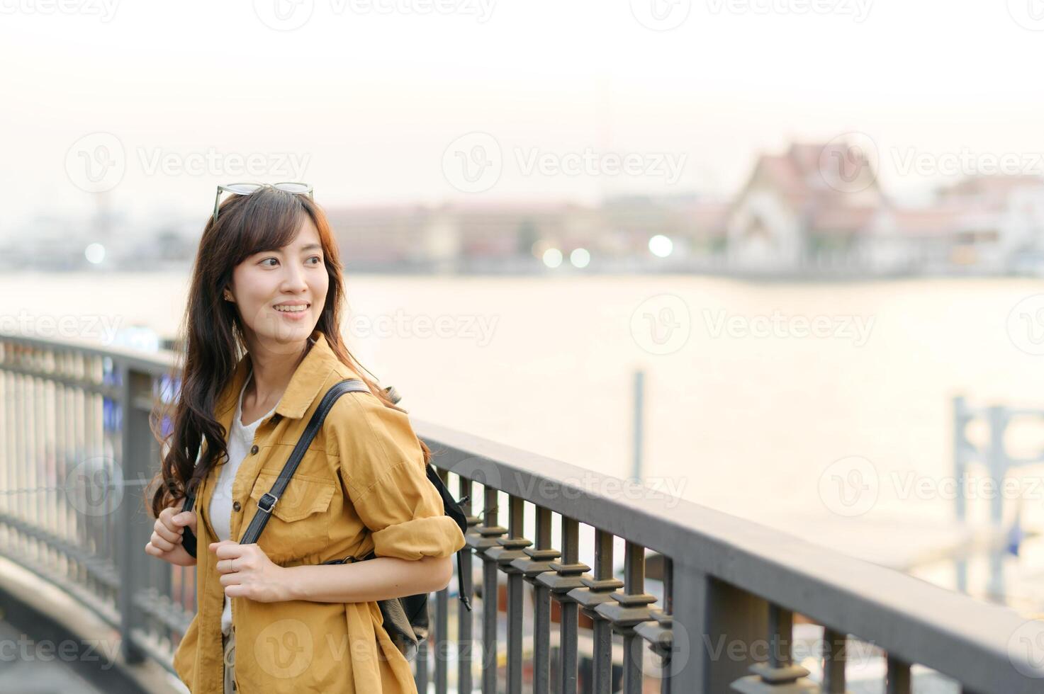 cheerful Asian woman with backpack enjoys serene riverside view during sunset, standing by railing with soft smile photo