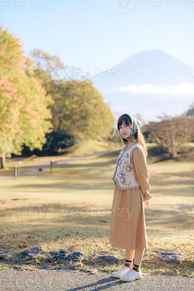 Asian woman stands gracefully in serene park with autumn foliage and mountain in background, exuding calm and elegance photo