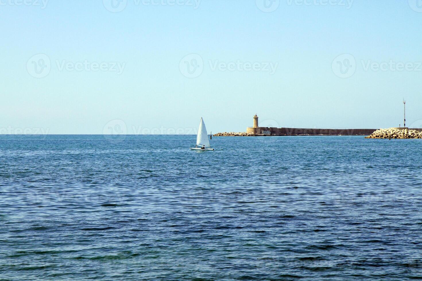 A sailboat in the open sea near a lighthouse and breakwater on a sunny day. photo