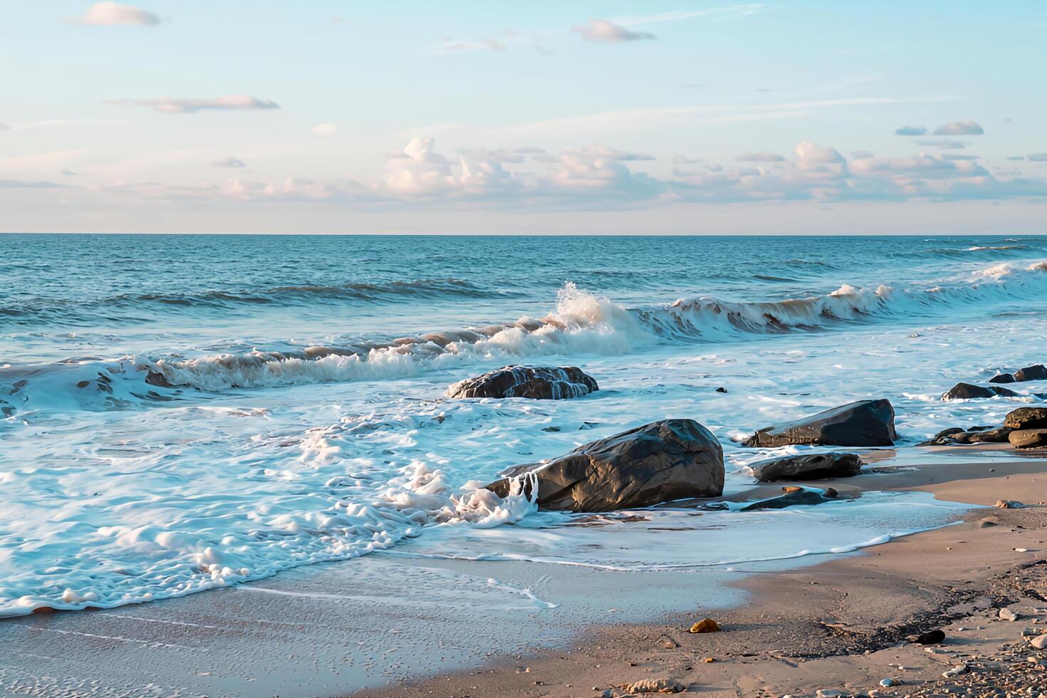Serene ocean waves gently crashing on sandy shore with dark rocks under a soft blue sky photo