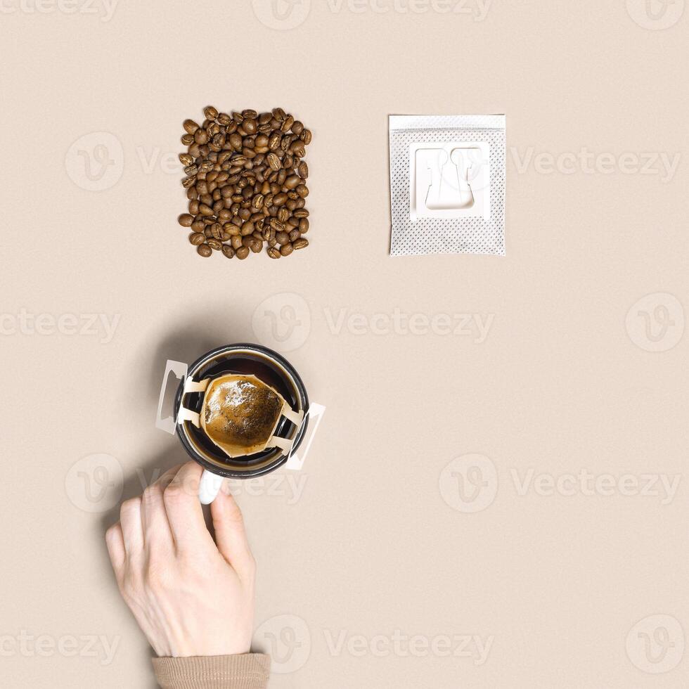 Woman hand hold cup and make black coffee drink in cup, drip coffee bag with ground coffee brewed in cup, paper bag drip coffee filter, beige background, top view, minimal flat lay photo